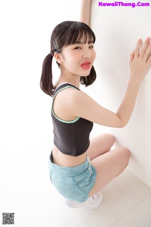 A woman in a black top and blue shorts leaning against a wall.