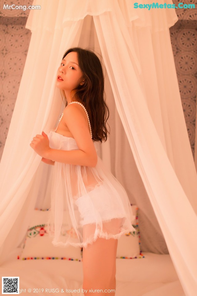 A woman in a white lingerie standing in front of a bed.